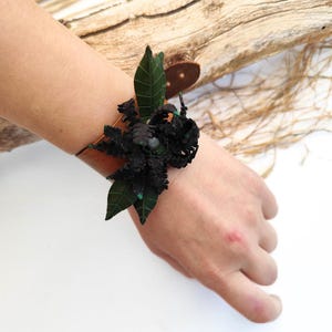 Könnte beinhalten: Ein braunes Lederarmband mit einem dunkelgrünen und schwarzen Blumendesign. Das Armband hat dunkelgrüne Blätter und eine schwarze, strukturierte Blume. Ein kleiner, brauner Knopf sichert das Armband.