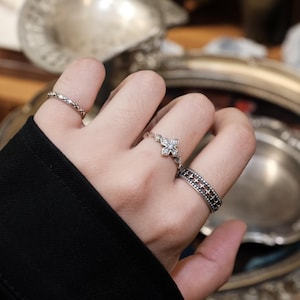 Könnte beinhalten: Eine Hand mit drei silbernen Ringen. Ein Ring hat ein Kreuzmuster, ein anderer ein sich wiederholendes Muster und der dritte ein filigranes Kettendesign. Die Ringe sind vor einem verschwommenen Hintergrund aus silbernen Servierteilen platziert.