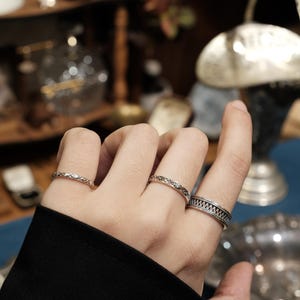 May include: Three silver-toned rings worn on three fingers. The ring on the index finger features a wide band with a textured, geometric pattern. The middle finger ring has a delicate, openwork design, and the ring finger band is thin and textured.