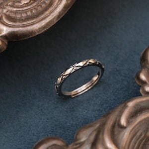 May include: A silver ring featuring a geometric diamond and dot pattern. The adjustable ring is set against a dark blue backdrop, with antique-style metalwork in the background.