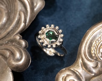 Pode incluir: Um anel de prata com uma pedra preciosa verde esmeralda central, rodeada por uma cravação decorativa em forma de raio de sol. O anel está sobre um fundo azul escuro, com peças de servir de prata ornamentadas de cada lado.