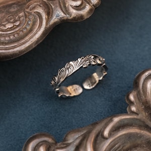 May include: A silver ring with an open design, adorned with an elaborate floral pattern. The ring is set against a dark blue backdrop, with decorative silver frames in the foreground and background. The ring's intricate details are highlighted.
