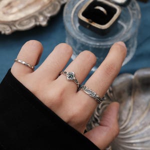 May include: A hand adorned with multiple silver rings, including one with a small green gem and others with detailed floral and geometric patterns. The rings are set against a backdrop of vintage-style decorative items.