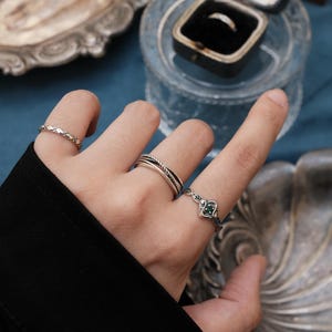 May include: A hand adorned with various silver rings, including a braided band, a ring with a green stone, and a plain band. A ring box with a ring inside is in the background, alongside a decorative silver dish. The rings are displayed on a blue surface.