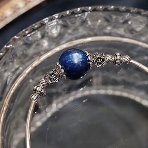 Könnte beinhalten: Ein silbernes Armreif-Armband mit einer großen, runden, tiefblauen Perle in der Mitte. Das Armband hat dekorative silberne Blumen- und Perlenakzente auf beiden Seiten der Perle. Das Armband wird in einer durchsichtigen Glasschale präsentiert.