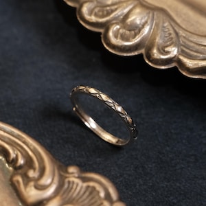 Puede incluir: Un anillo dorado con un patrón de diamantes repetido. El anillo está sobre un fondo oscuro, con elementos decorativos dorados ornamentados en primer plano y en segundo plano. El anillo es una joya delicada.
