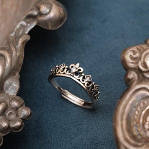 May include: A silver-colored crown ring with a fleur-de-lis design and small, clear gemstones. The ring is adjustable and sits on a blue surface, with ornate silver objects in the background. The ring is a piece of jewelry.
