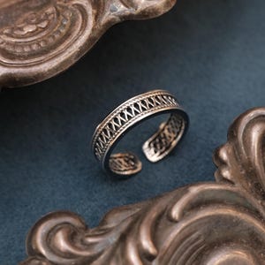 May include: Silver adjustable ring with an openwork design. The ring has a detailed pattern around the band. It is displayed on a dark blue surface, with a decorative, ornate tray in the background.