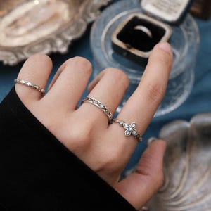 Puede incluir: Primer plano de tres anillos de plata con diseños intrincados. Uno presenta un patrón de diamantes, otro un diseño geométrico repetido y el tercero tiene forma de cruz con pequeñas piedras preciosas. Un joyero se ve en el fondo.