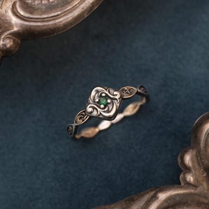 May include: A silver ring with an emerald-green gemstone set in a square, ornate setting. The band features a delicate, swirling design. The ring is displayed on a dark blue surface, framed by a decorative silver border.