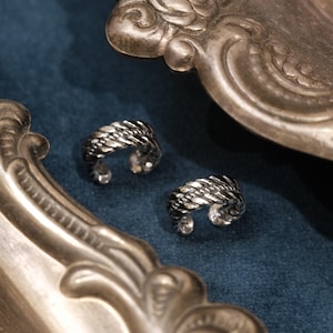 Puede incluir: Un par de pendientes tipo ear cuff plateados con una textura trenzada similar a una cuerda. Cada pieza es un aro circular abierto con un acabado metálico pulido, que muestra un patrón trenzado detallado en toda su circunferencia.