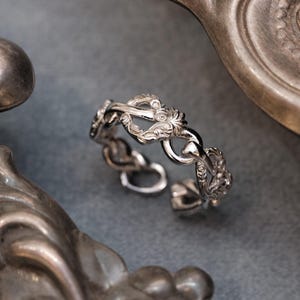May include: A silver ring with an intricate chain-link design. The ring showcases detailed floral and scrollwork patterns. It is set against a muted grey background with antique silverware.