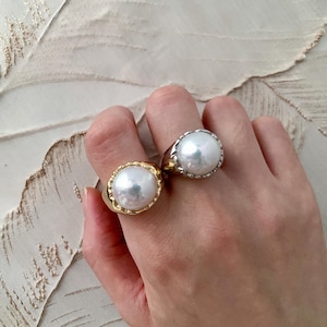 May include: Two rings featuring large, round, white pearls set in metal bands. One ring has a polished gold-tone band with a textured, scalloped edge around the pearl, while the other ring features a smooth, silver-tone band with a scalloped setting.