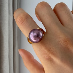 May include: A gold ring featuring a large, round, lustrous purple pearl as the centerpiece. The band is crafted from polished yellow gold and includes small, clear accent stones set into the metal on either side of the central pearl setting.