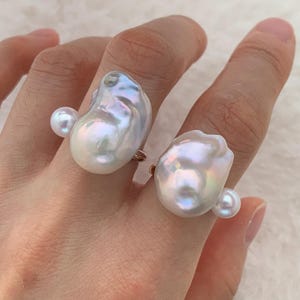 May include: Two rings with large, irregularly shaped white pearls. Each ring features a smaller, round pearl accent. The rings have gold-colored bands and are displayed on a hand.