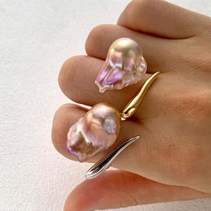 May include: A distinctive ring with two large, iridescent baroque pearls. The ring has a gold and silver metal design that curves around the pearls. The pearls are pink and cream-coloured with a textured surface.