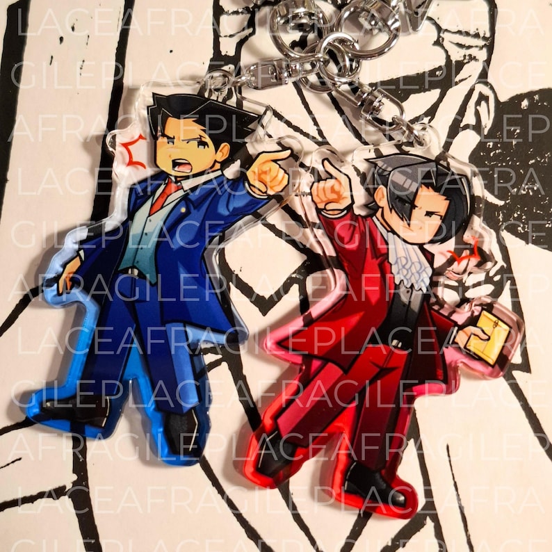 May include: Two clear acrylic keychains featuring anime-style characters. One character wears a blue suit and points forward, while the other wears a red suit with a white cravat, points forward, and holds a small yellow book. Both have metal clasps.
