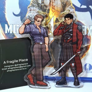 May include: Two acrylic keychains featuring anime-style male characters. One character wears a purple shirt and sunglasses, the other a red and black outfit and holds a sword. The keychains are displayed in front of a promotional card with the text "A Fragile Place".
