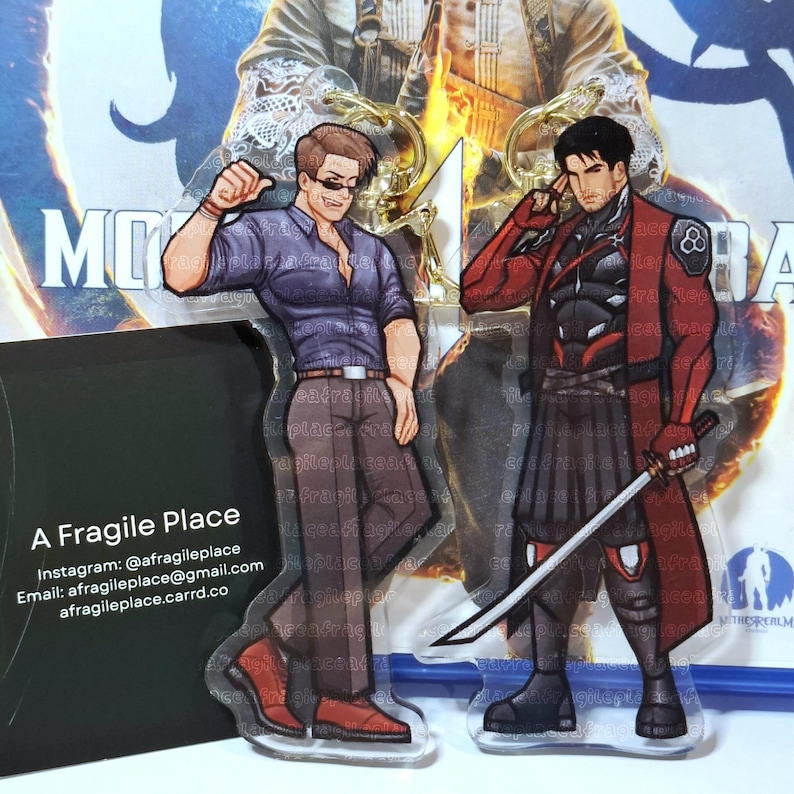 May include: Two acrylic keychains featuring anime-style male characters. One character wears a purple shirt and sunglasses, the other a red and black outfit and holds a sword. The keychains are displayed in front of a promotional card with the text "A Fragile Place".