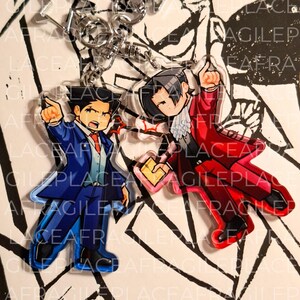 May include: Two acrylic keychains featuring chibi-style characters in suits. One character wears a blue suit and points forward, while the other wears a red suit with a white cravat, holds a yellow folder, and points upward. Both have silver clasps.