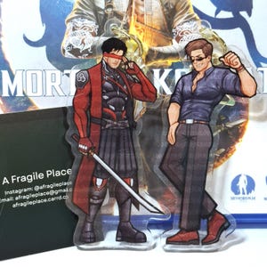 Peut inclure: Deux figurines acryliques de personnages de Mortal Kombat. L'une porte une tenue rouge et noire avec une épée, l'autre une chemise et un pantalon bleu foncé. Le fond comprend le texte "MORTAL KOMBAT".