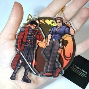 May include: Two acrylic keychains featuring male characters in action poses. One keychain depicts a character in a red and black outfit with a sword. The other shows a character in a blue shirt and brown pants. Both have gold keychains.