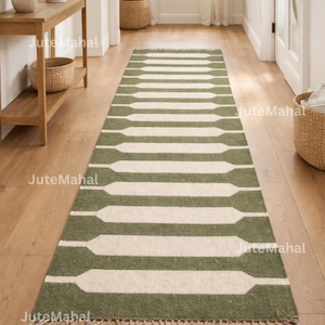 May include: A green and white patterned runner rug with a rectangular design. The rug has a series of green rectangular shapes with white spaces in between. The rug has a fringe on the bottom edge. The rug is on a wooden floor.