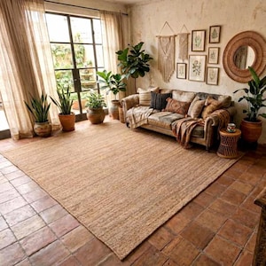 May include: A large, rectangular jute rug in a living room. The rug is a natural tan colour with a woven texture. The room features a sofa with decorative cushions, potted plants, and a large window with sheer curtains. The floor is tiled.