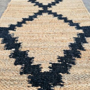 May include: A rectangular jute rug with a geometric black pattern. The rug has a natural beige background and a repeating diamond-like design in black. The rug is made of woven fibers and is suitable for indoor use.