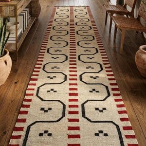 May include: A long, rectangular rug with a cream-colored background and a repeating pattern of black and red geometric shapes. The rug has a fringe on both ends and is placed on a wooden floor. The design is symmetrical and modern.