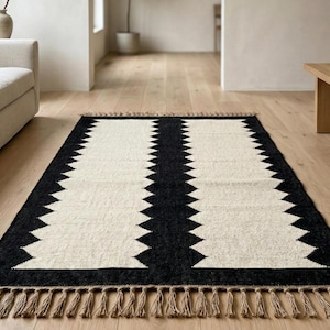 May include: A rectangular rug with a black border and a cream-colored central design. The design features two vertical columns of zig-zag patterns. The rug has a fringe on both ends, adding a decorative element. This rug is suitable for living rooms and bedrooms.