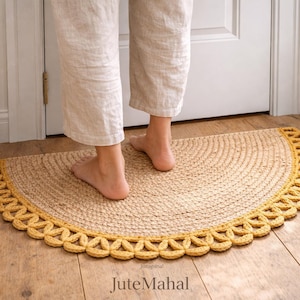 May include: A semi-circular rug made of natural tan woven fibers featuring a decorative mustard yellow crocheted border with a repeating loop pattern. The rug is positioned on a wooden floor and displays the text JuteMahal at the bottom center.