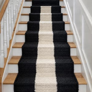 May include: A black and cream-colored stair runner with a central cream stripe. The runner is installed on wooden stairs with white risers and a white banister. The runner adds a modern touch to the staircase.