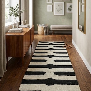 May include: A long rectangular runner rug featuring a repeating geometric pattern of bold black horizontal bars against a cream-colored background, finished with a natural tan woven border at the short ends, laid flat on a dark wooden floor.
