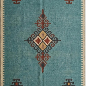 May include: A rectangular area rug in teal blue featuring a central diamond-shaped geometric pattern in rust, cream, and dark brown. The rug has a cream-colored border, four small matching geometric motifs in the corners, and fringed edges.