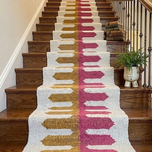 May include: A stair runner with a white background and a design of mustard yellow and magenta shapes. The runner is installed on wooden stairs with a wooden handrail and a decorative plant in a pot.