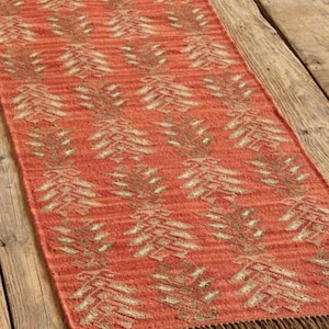 Könnte beinhalten: Ein rechteckiger, orange-roter Teppich mit einem sich wiederholenden Muster aus stilisierten, hellbeige baumartigen Formen. Der Teppich hat einen braunen Fransen an den kurzen Enden. Der Teppich liegt auf einem Holzboden.