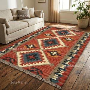 May include: A rectangular red rug with geometric patterns in blue, cream, and yellow. The rug has a fringed edge and is placed on a wooden floor. The rug is in a living room setting, near a beige sofa.