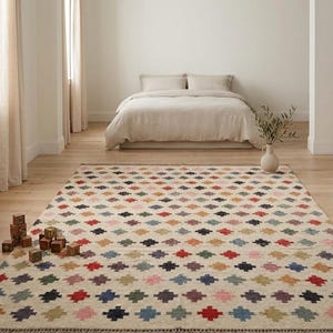 May include: A colorful area rug with a geometric pattern of diamonds in various colors, including red, blue, and yellow. The rug is placed in a bedroom with a bed and wooden floor. The rug has a fringed edge.