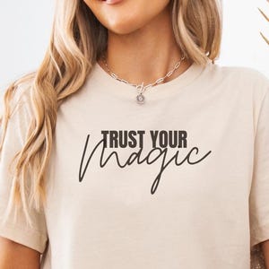 May include: A beige short-sleeve t-shirt featuring the black text "TRUST YOUR MAGIC" printed on the chest. The shirt has a crew neckline and a standard fit, paired with light blue denim shorts and a textured beige cardigan draped over the arms.