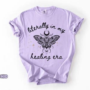 May include: A light purple t-shirt featuring a black graphic of a moth with a skull pattern, a crescent moon, and small gold stars. The text "literally in my healing era" is printed in black script above and below the moth illustration.
