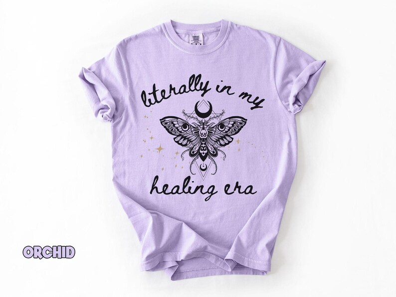 May include: A light purple t-shirt featuring a black graphic of a moth with a skull pattern, a crescent moon, and small gold stars. The text "literally in my healing era" is printed in black script above and below the moth illustration.