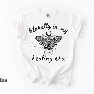 May include: A white t-shirt featuring a black graphic of a moth with a skull pattern, crescent moons, and geometric lines. The text "literally in my healing era" is printed in black script above and below the moth, with small gold stars.