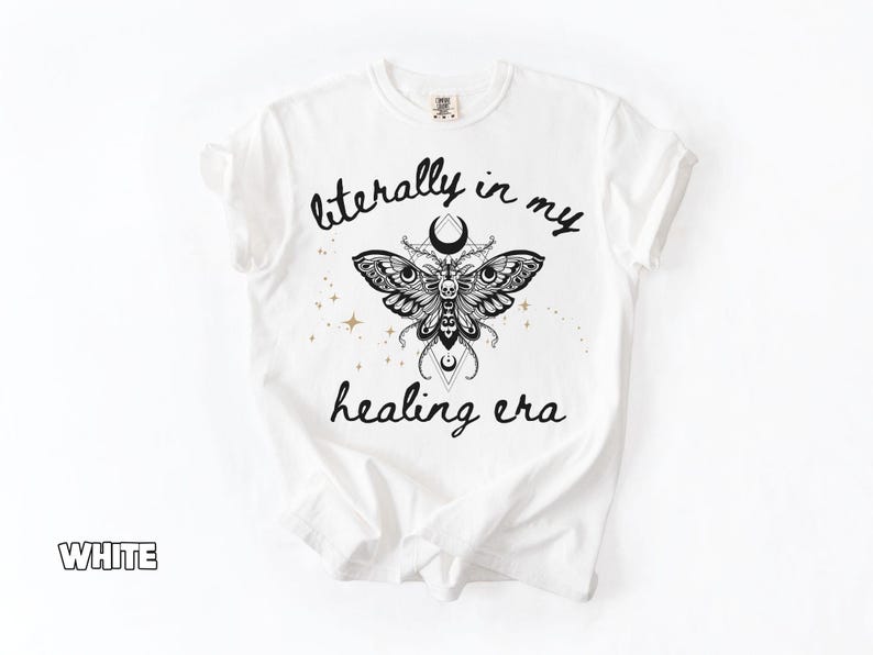 May include: A white t-shirt featuring a black graphic of a moth with a skull pattern, crescent moons, and geometric lines. The text "literally in my healing era" is printed in black script above and below the moth, with small gold stars.