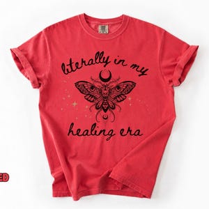 May include: A red short-sleeved t-shirt featuring a black graphic of a moth with intricate wing patterns, a crescent moon, and small stars. The text "literally in my healing era" is printed in a black script font above and below the moth.