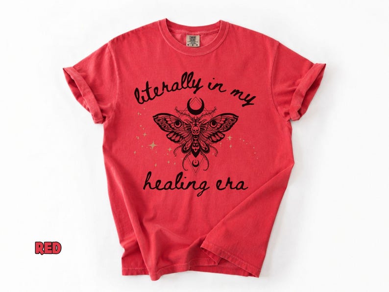 May include: A red short-sleeved t-shirt featuring a black graphic of a moth with intricate wing patterns, a crescent moon, and small stars. The text "literally in my healing era" is printed in a black script font above and below the moth.