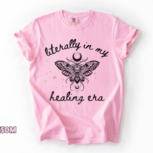 May include: A light pink t-shirt featuring a black graphic of a moth with intricate wing patterns, a crescent moon, and geometric shapes. The text "literally in my healing era" is printed in black script above and below the moth illustration.