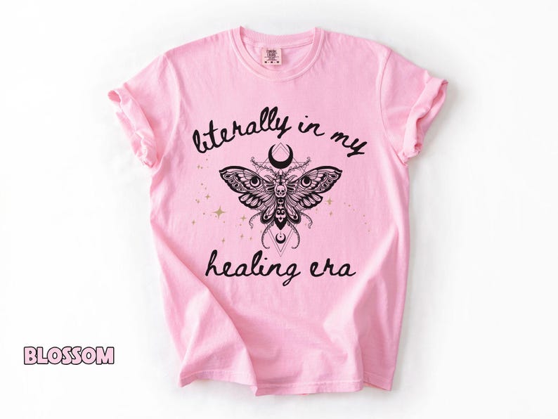 May include: A light pink t-shirt featuring a black graphic of a moth with intricate wing patterns, a crescent moon, and geometric shapes. The text "literally in my healing era" is printed in black script above and below the moth illustration.