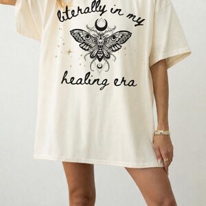 May include: An off-white short-sleeve t-shirt featuring a black graphic of a moth with a skull and crescent moon. The text "literally in my healing era" is printed in a black script font above and below the moth, surrounded by small stars.
