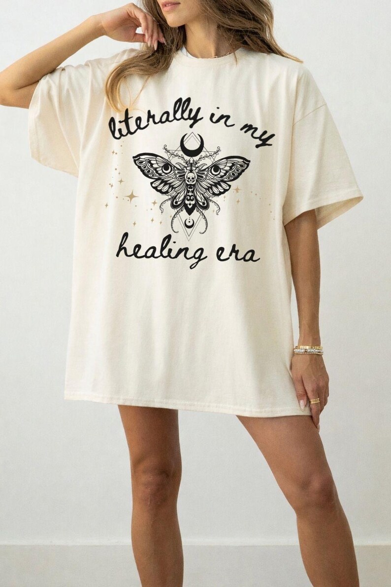 May include: An off-white short-sleeve t-shirt featuring a black graphic of a moth with a skull and crescent moon. The text "literally in my healing era" is printed in a black script font above and below the moth, surrounded by small stars.